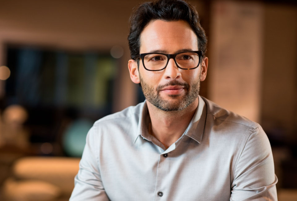 Man with glasses and a beard, wearing a light blue shirt, sitting in a modern setting, representing eye care and vision health at Henry Eye Clinic.
