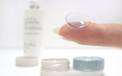 Contact lens resting on a fingertip with contact lens case and solution bottle in the background, illustrating safe contact lens handling and care.