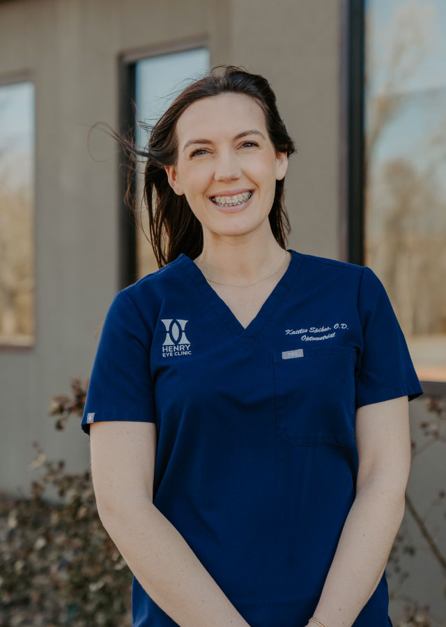 Kaitlin N. Spikes, O.D., in blue scrubs with "Henry Eye Clinic" logo, smiling outdoors, representing eye care expertise.